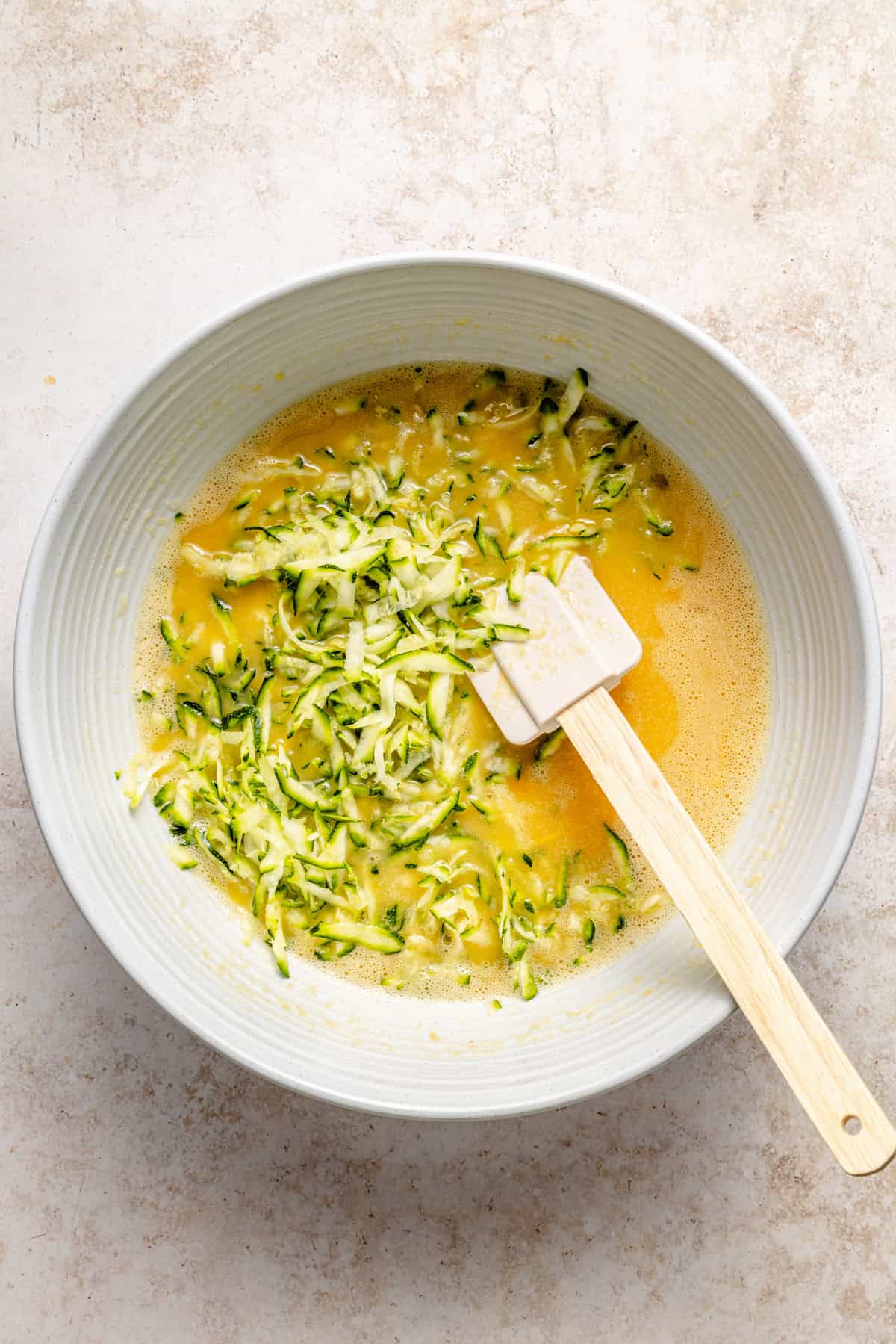 A white bowl with whisked eggs and shredded zucchini being mixed with a white spatula on a light countertop—a fresh start for paleo zucchini bread.