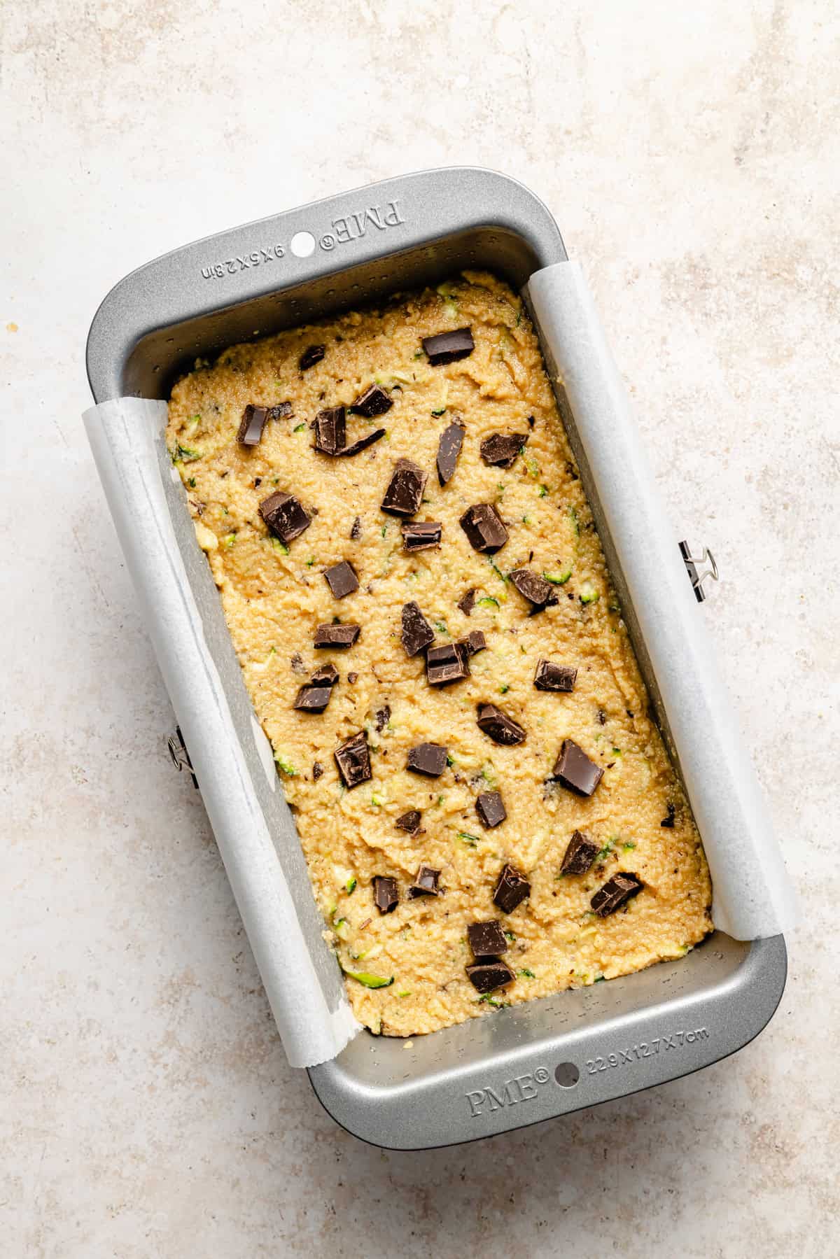 A loaf pan lined with parchment paper holds unbaked paleo zucchini bread batter topped with dark chocolate chunks, ready to be baked. The batter features green flecks of zucchini and rests on a light-colored surface.