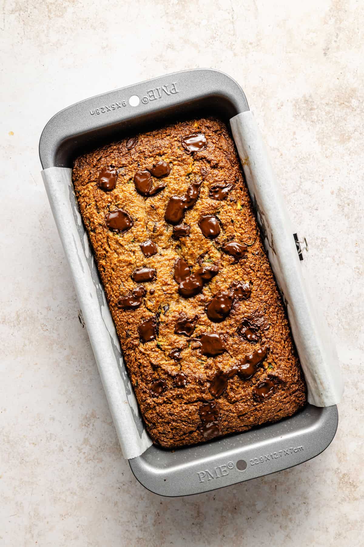 A freshly baked loaf of paleo zucchini bread with melted chocolate chunks on top sits in a rectangular metal baking pan lined with parchment paper on a light countertop.