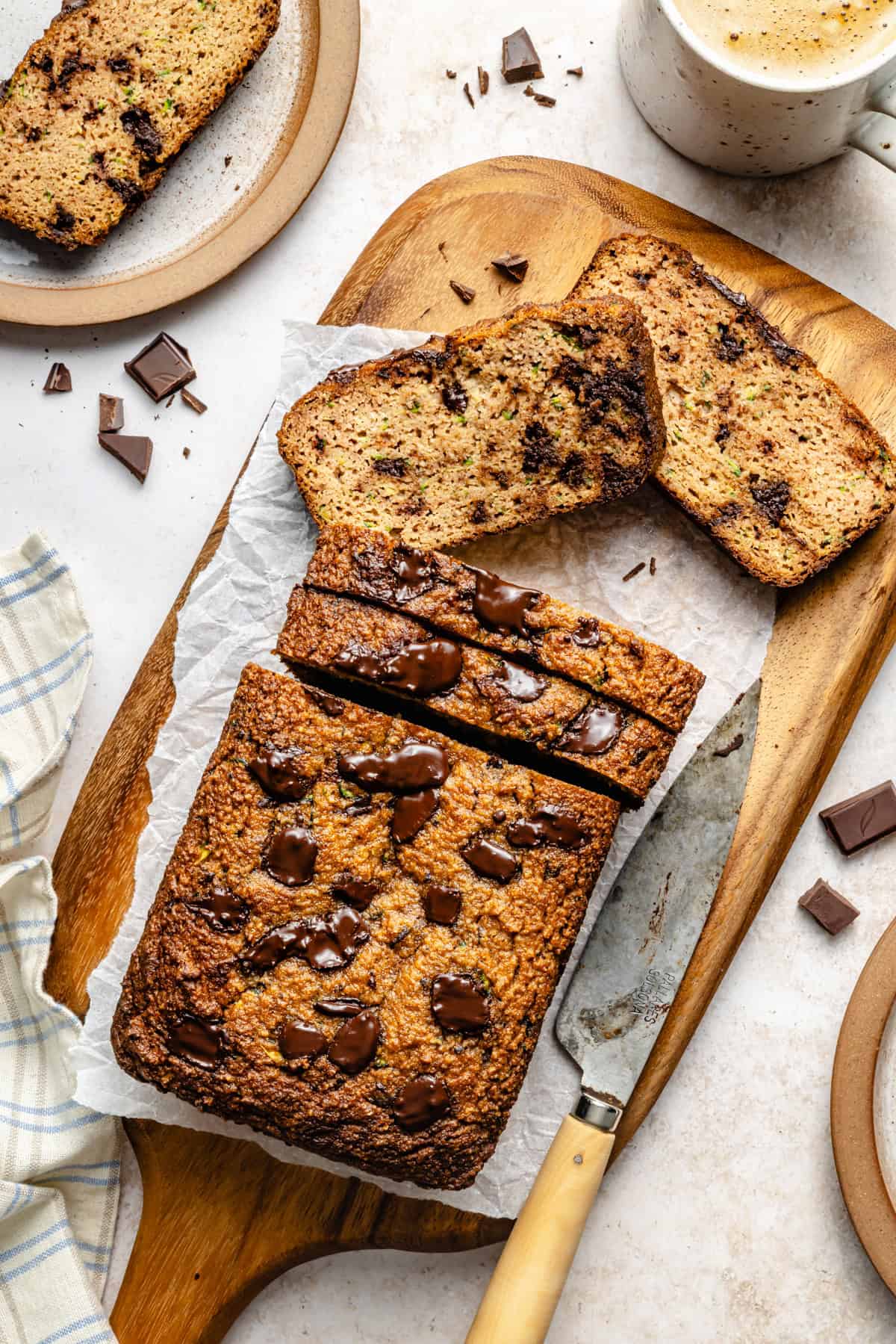 A sliced loaf of chocolate chip banana bread rests on a wooden board with a knife beside it. Two slices are placed near the loaf, along with chocolate chunks and a mug of coffee—perfect inspiration for your next paleo zucchini bread. A beige napkin sits to the side.