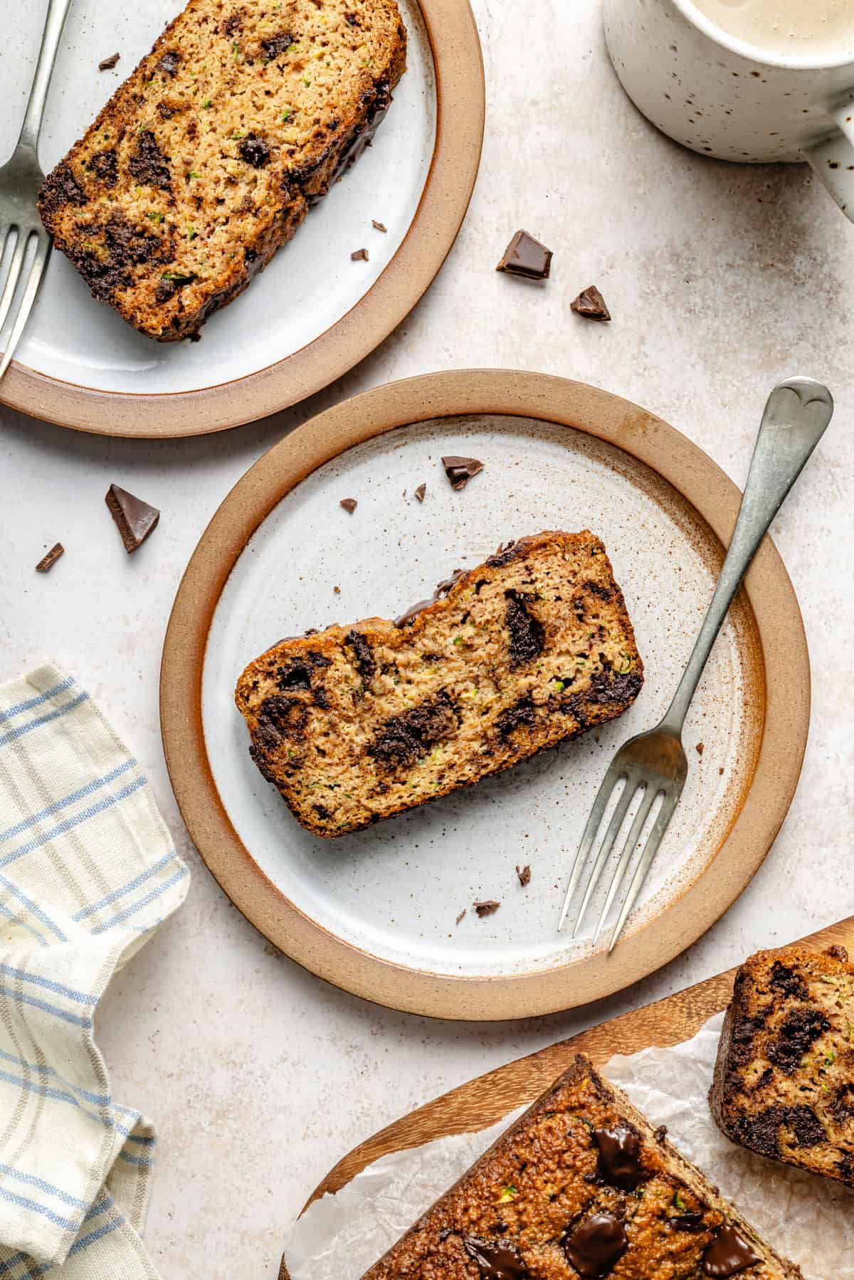 Two plates with slices of paleo zucchini bread, each with a fork. A cup of coffee sits nearby, along with a checkered napkin and scattered chocolate pieces on a light table.