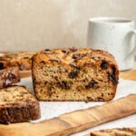 A sliced loaf of paleo zucchini bread with chocolate chunks sits on parchment paper atop a wooden board, with a white ceramic mug in the background.