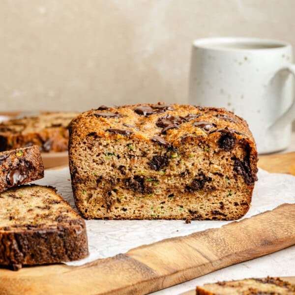 A sliced loaf of paleo zucchini bread with chocolate chunks sits on parchment paper atop a wooden board, with a white ceramic mug in the background.