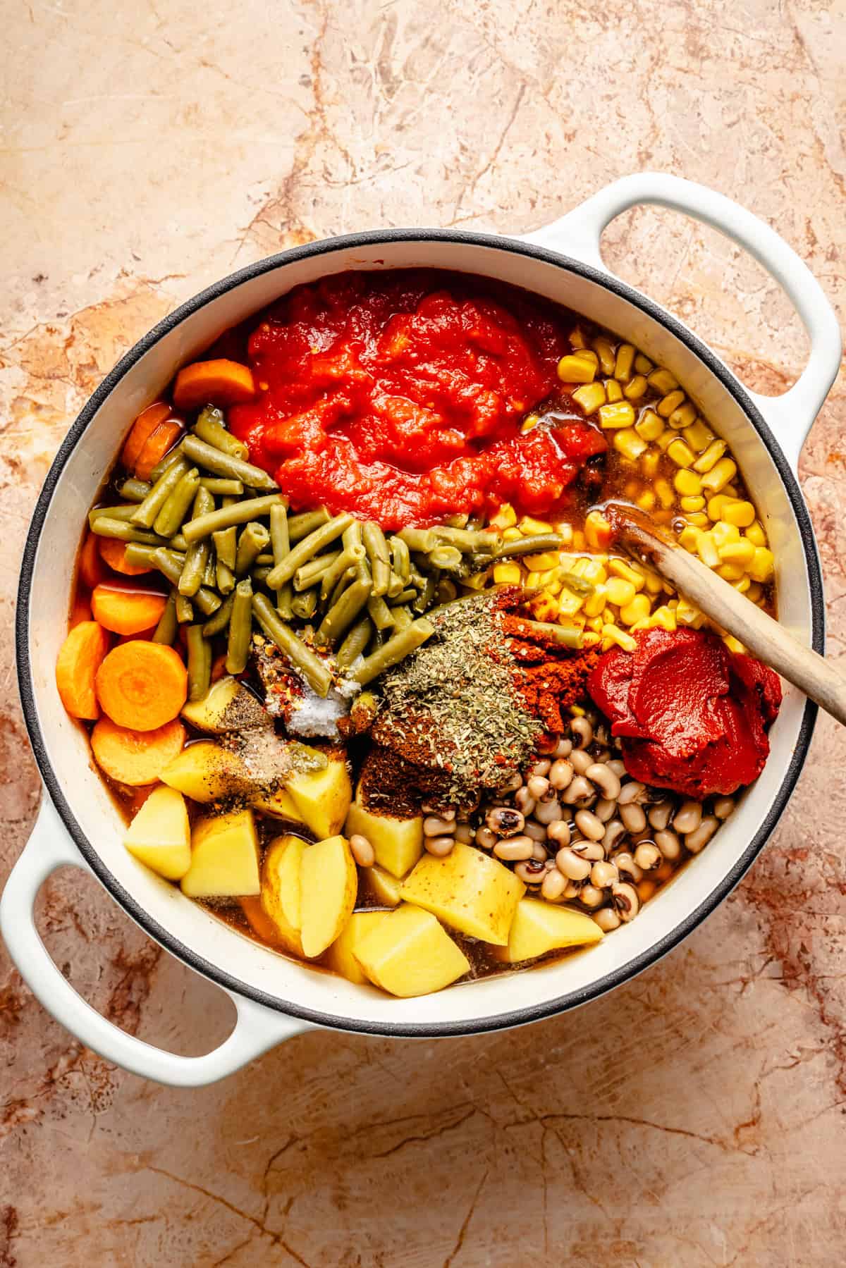 A white pot filled with chopped potatoes, carrots, green beans, corn, black-eyed peas, diced tomatoes, tomato paste, and assorted spices sits ready to be mixed for a hearty cowboy soup stew. A wooden spoon rests inside the pot.