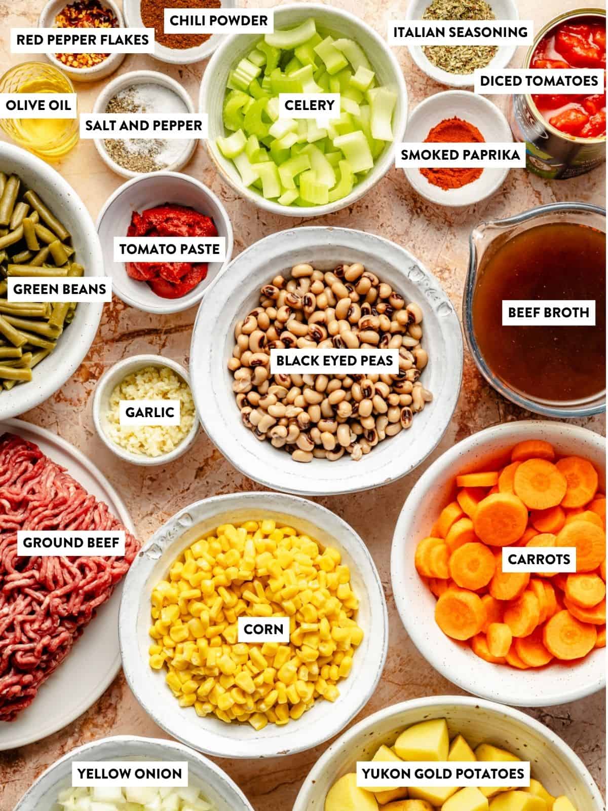 Various labeled ingredients for cowboy soup, including black-eyed peas, ground beef, corn, carrots, celery, yellow onion, Yukon Gold potatoes, beef broth, diced tomatoes, tomato paste, garlic, and multiple seasonings.