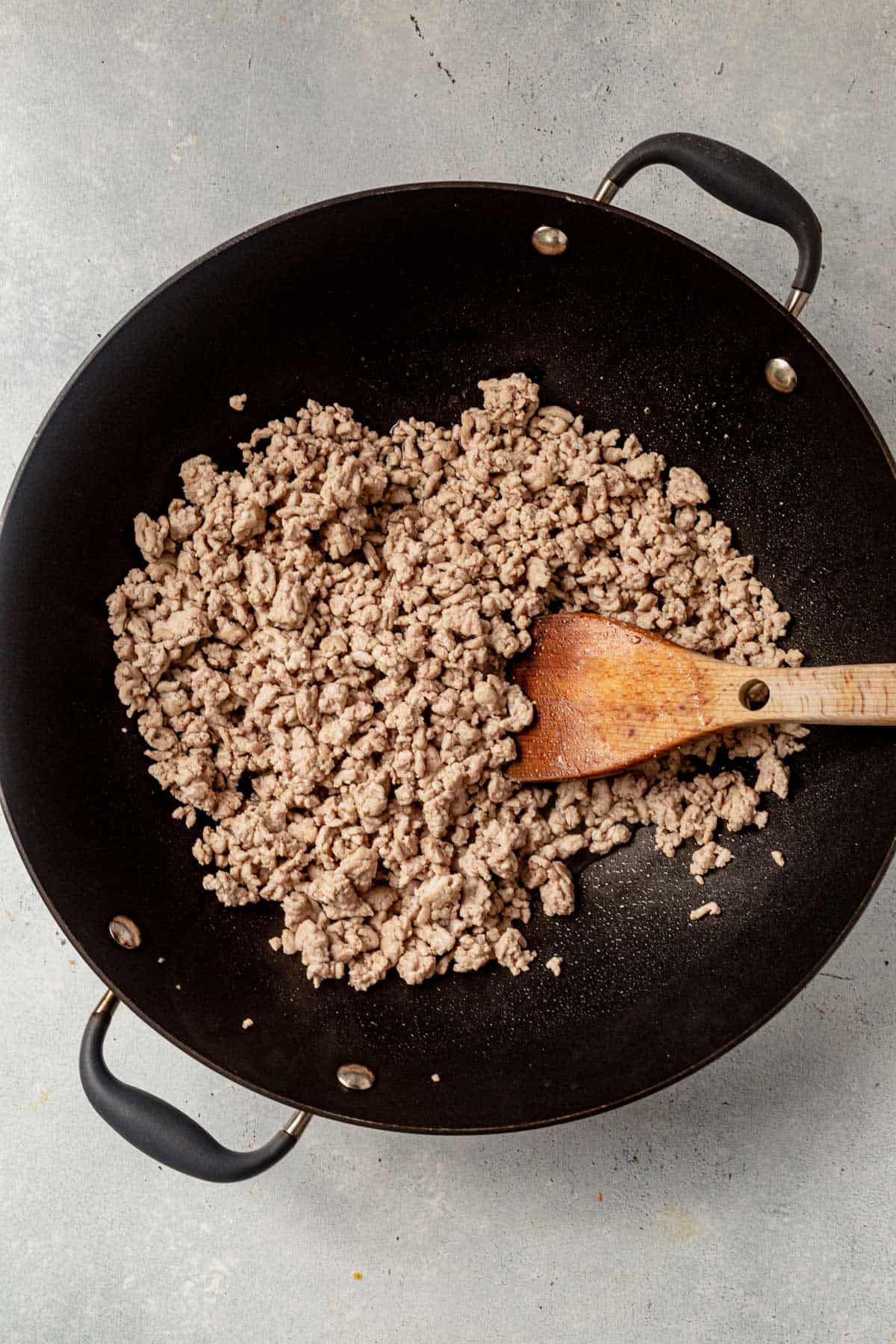 browning ground pork in a wok with a wooden spoon.