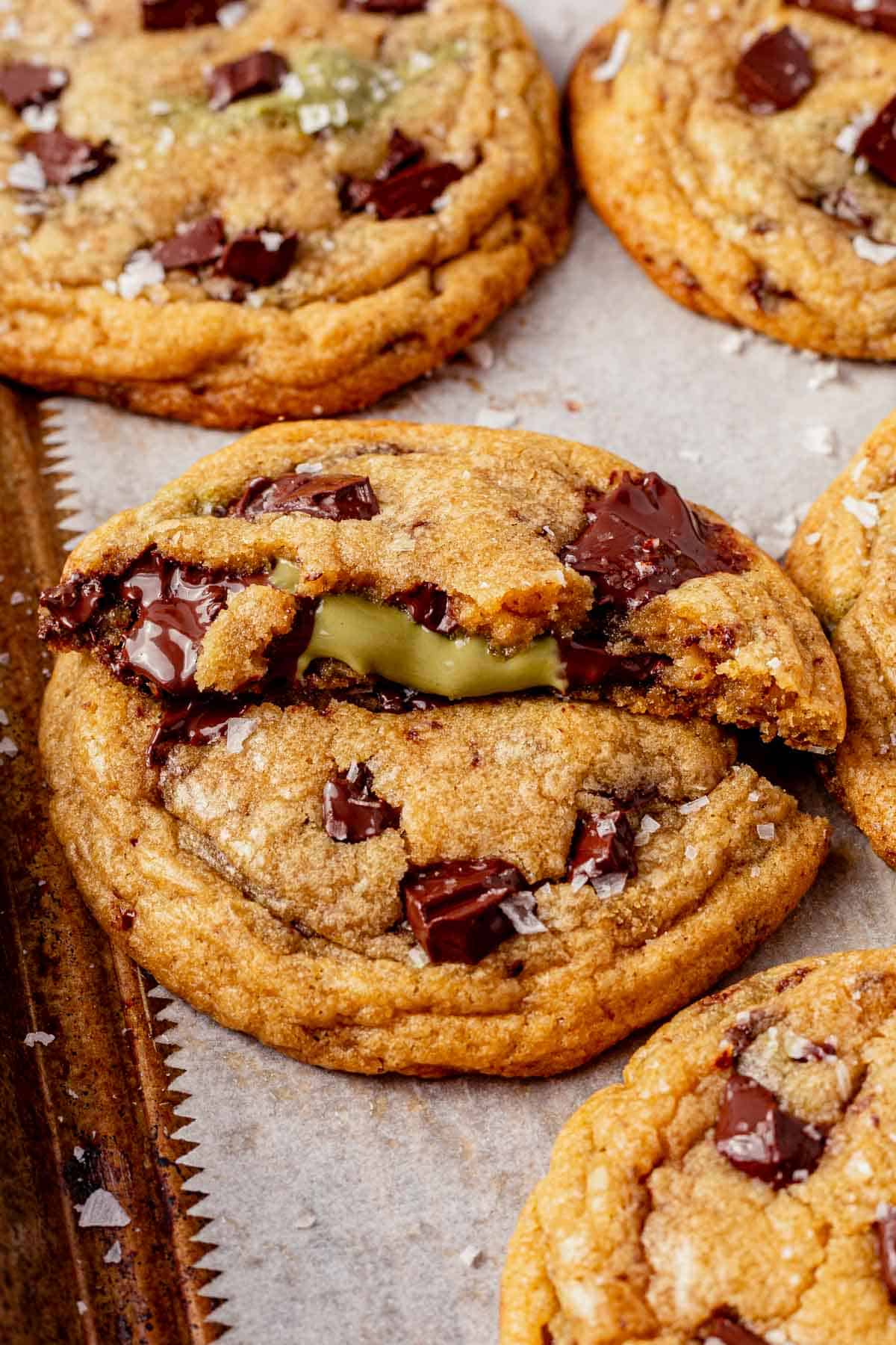 a brown butter pistachio chocolate chip cookie broken in half on a cookie sheet.