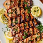 Grilled salmon filets with charred grill marks, garnished with fresh parsley and lemon slices on a white serving platter.