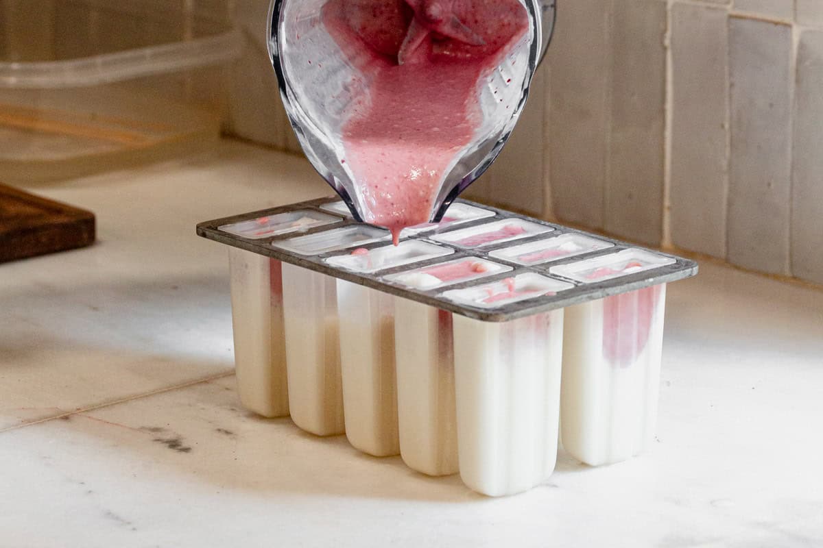 pouring strawberry filling for ice cream bars on top of the vanilla layer in popsicle molds.