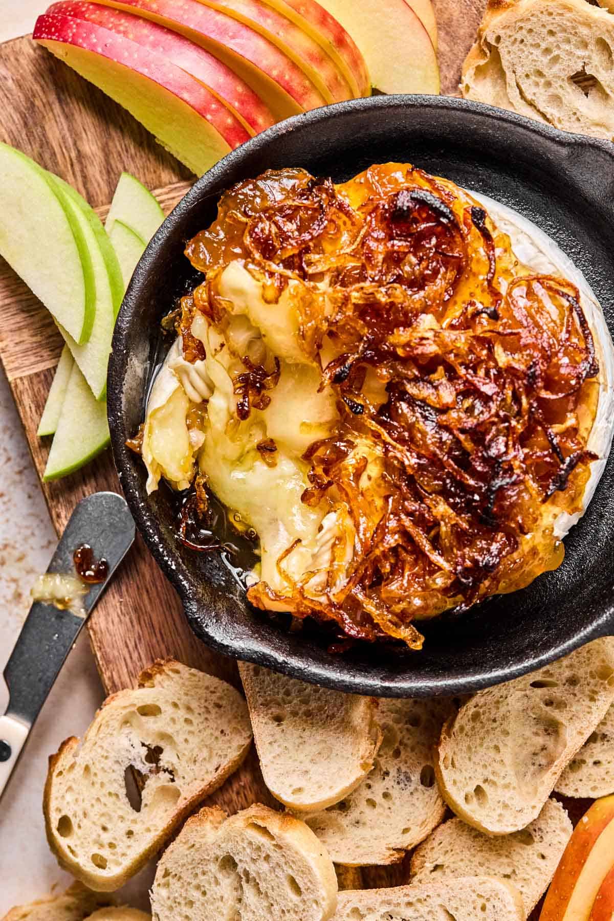 baked brie with fig jam and caramelized onions on top sitting in a small cast iron with a cheese spreader, apple slices, and crostini around it