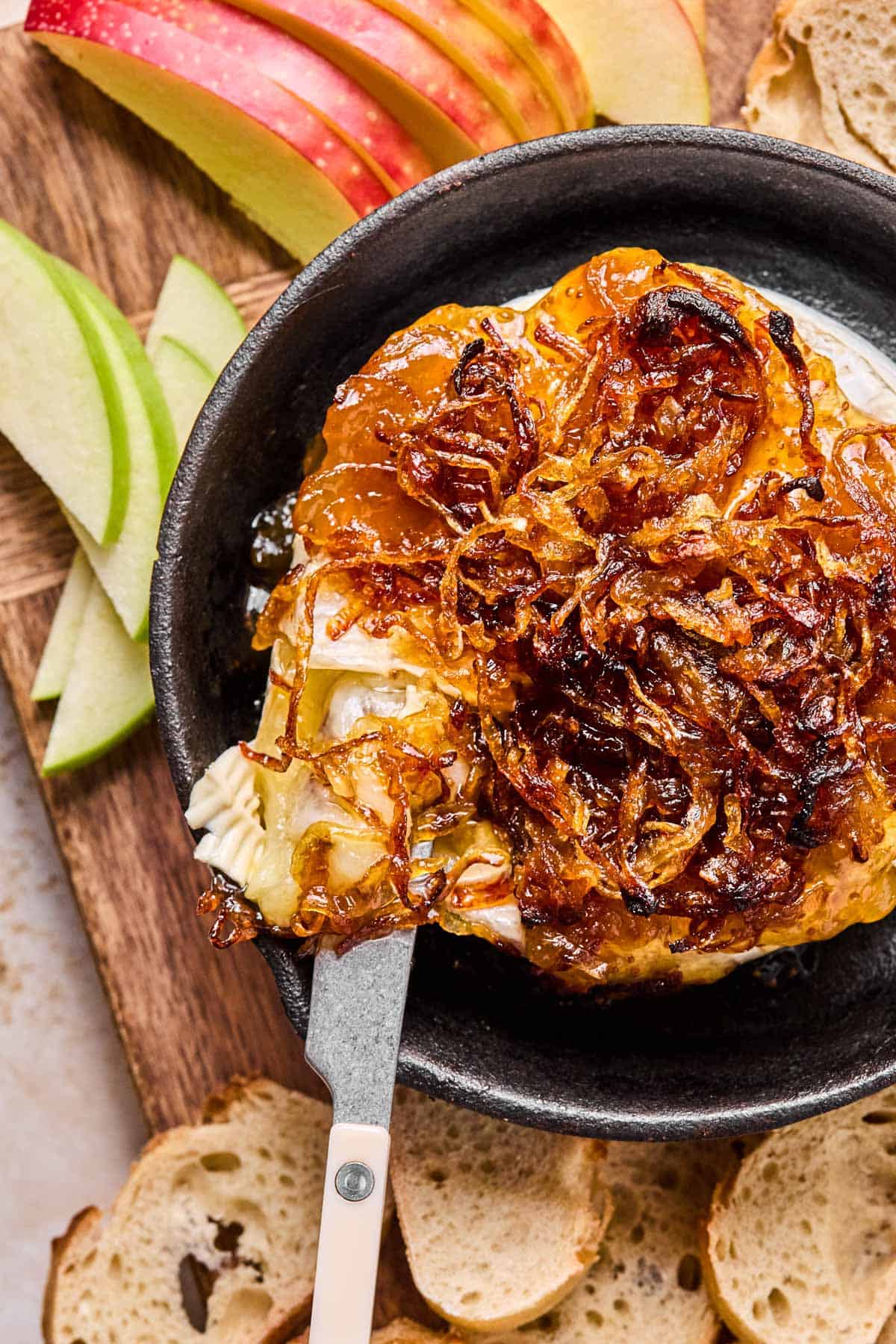 a cheese spreader dipped into a cast iron with melted wheel of brie covered in fig jam and caramelized onions