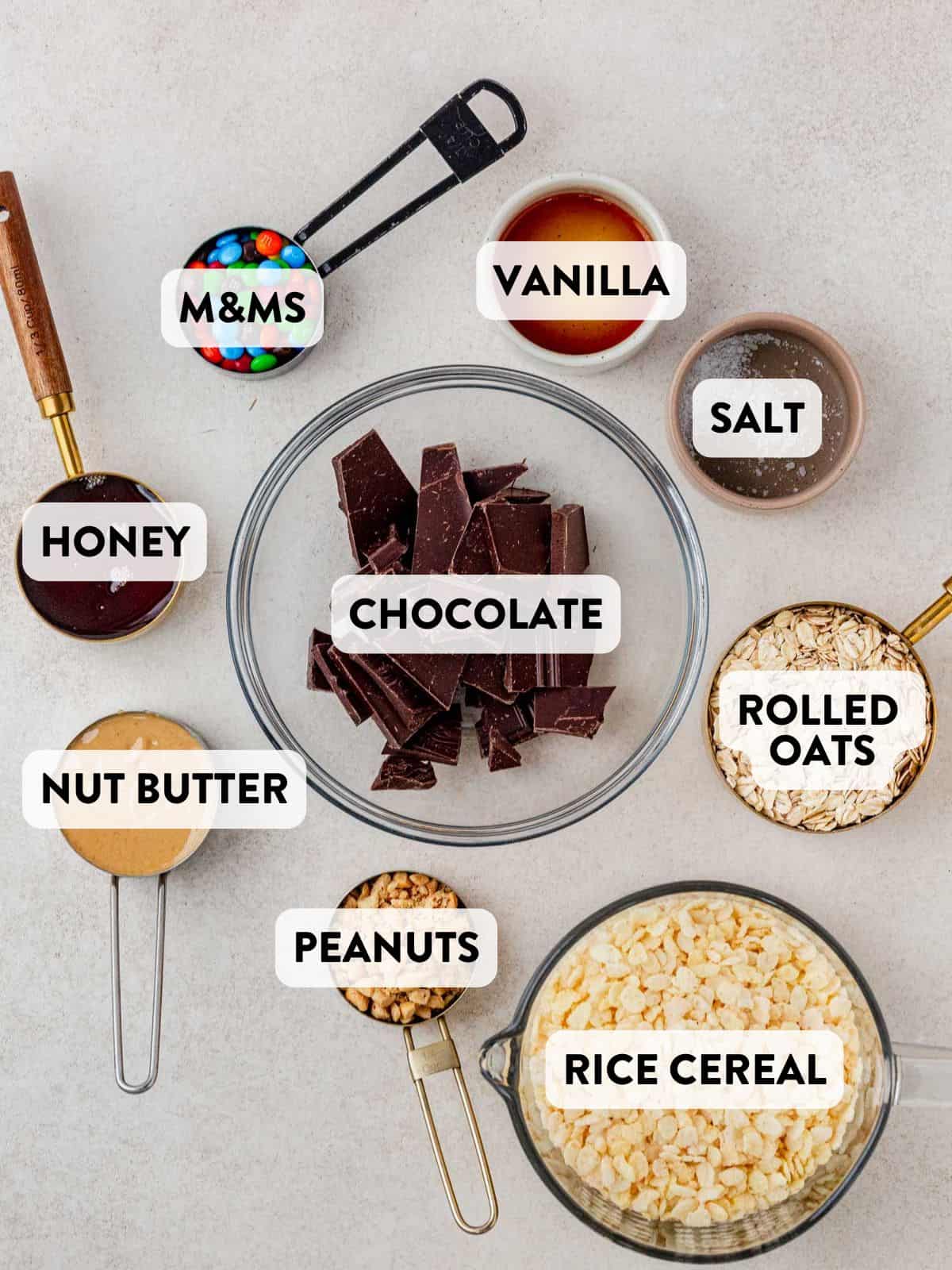 Flat lay of homemade Kudos granola bar ingredients on a light background, including chocolate chunks, M&Ms, vanilla extract, salt, honey, nut butter, peanuts, rolled oats, and rice cereal—all labeled for reference.
