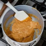 Nut butter and honey mixture bubbling and being stirred with a spatula in a white saucepan over a gas stovetop.