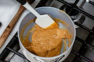 Nut butter and honey mixture bubbling and being stirred with a spatula in a white saucepan over a gas stovetop.
