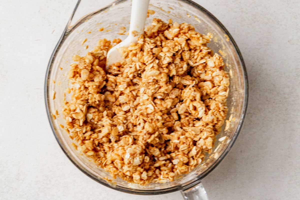 Rolled oats and rice cereal mixture coated in nut butter and honey, stirred together in a glass mixing bowl with a white spatula.