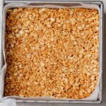 Granola bar mixture pressed firmly into a parchment-lined square baking pan, ready to chill in the fridge.