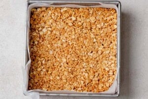 Granola bar mixture pressed firmly into a parchment-lined square baking pan, ready to chill in the fridge.