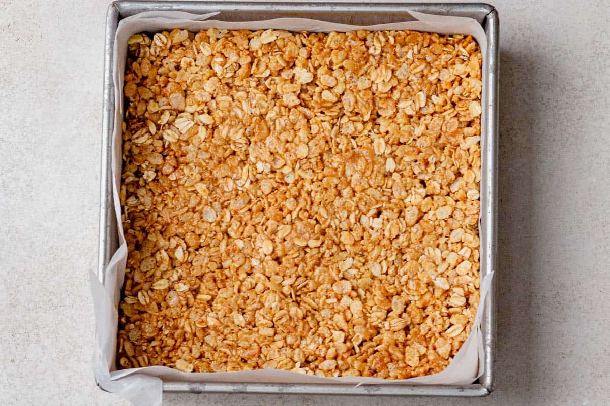 Granola bar mixture pressed firmly into a parchment-lined square baking pan, ready to chill in the fridge.