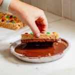Hand dipping a homemade copycat Kudos granola bar topped with mini M&Ms into melted chocolate for a fun and easy no-bake treat.