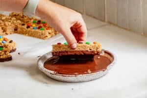Hand dipping a homemade copycat Kudos granola bar topped with mini M&Ms into melted chocolate for a fun and easy no-bake treat.
