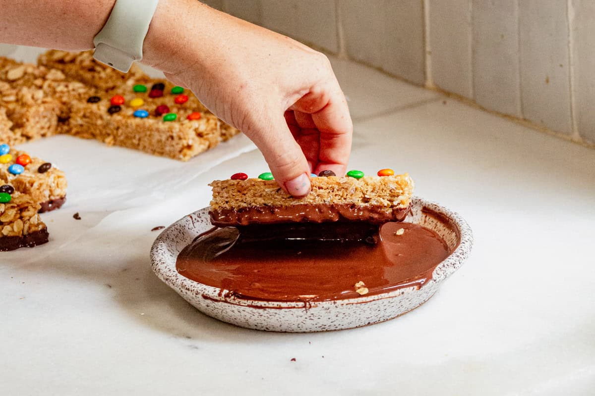 Hand dipping a homemade copycat Kudos granola bar topped with mini M&Ms into melted chocolate for a fun and easy no-bake treat.