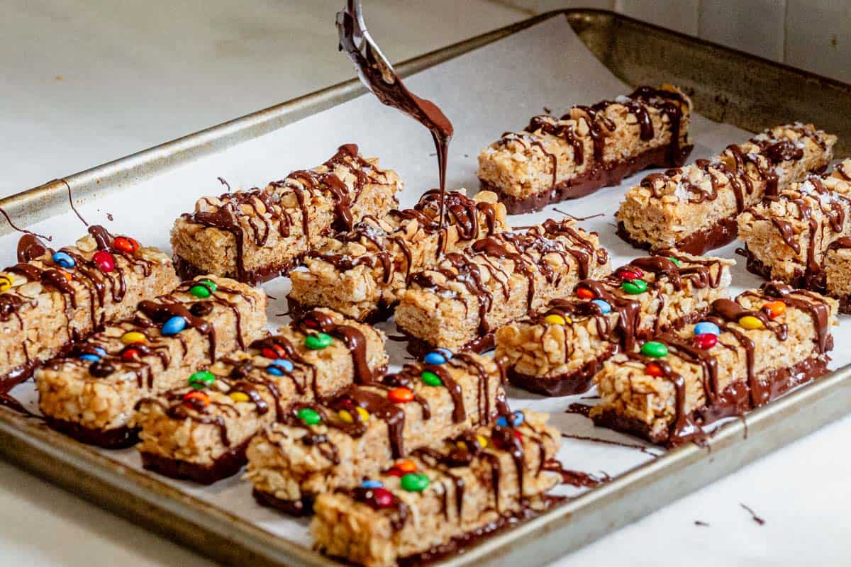 Tray of no-bake copycat Kudos bars with rice cereal, oats, and peanut butter being drizzled with melted chocolate.