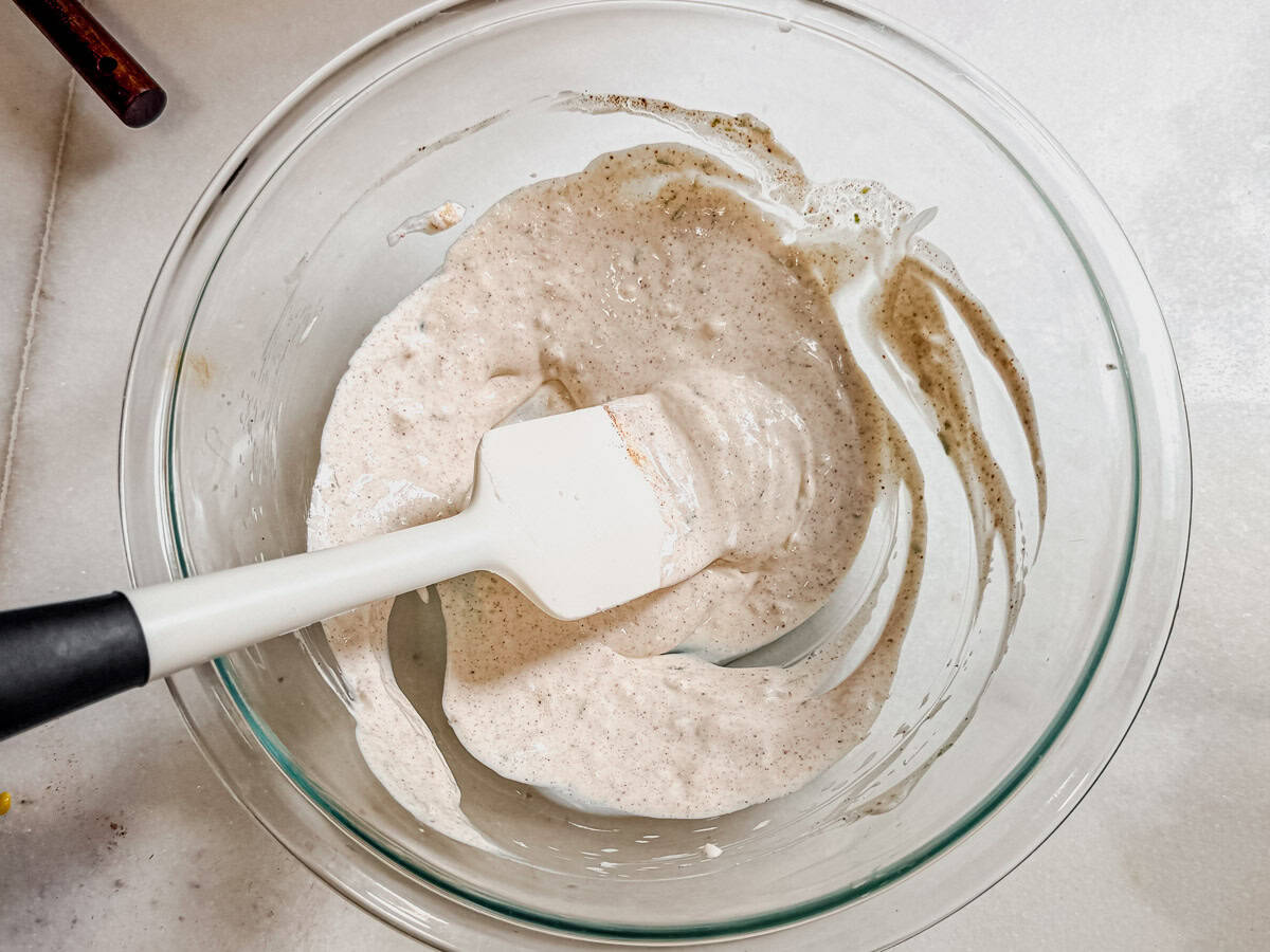 Glass mixing bowl with creamy lime dressing being stirred with a rubber spatula for Mexican street corn salad.