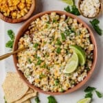 Bowl of Mexican street corn salad topped with lime wedges and fresh cilantro, surrounded by chips and extra grilled corn.
