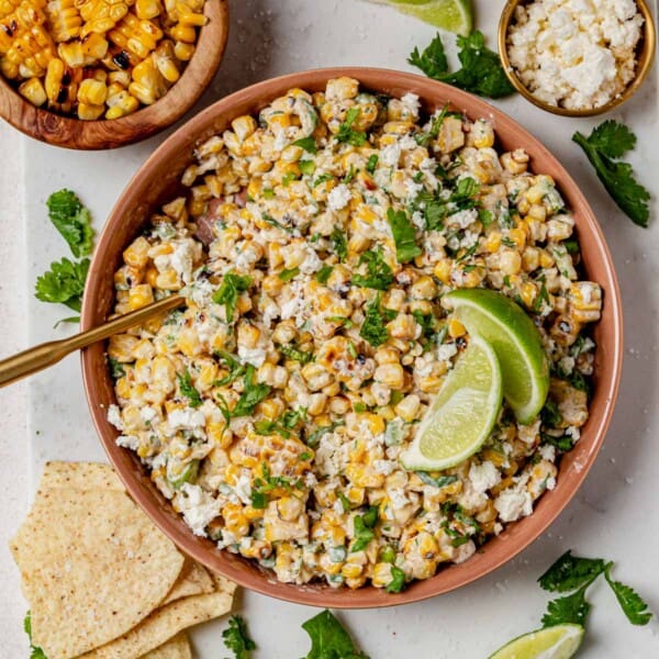 Bowl of Mexican street corn salad topped with lime wedges and fresh cilantro, surrounded by chips and extra grilled corn.