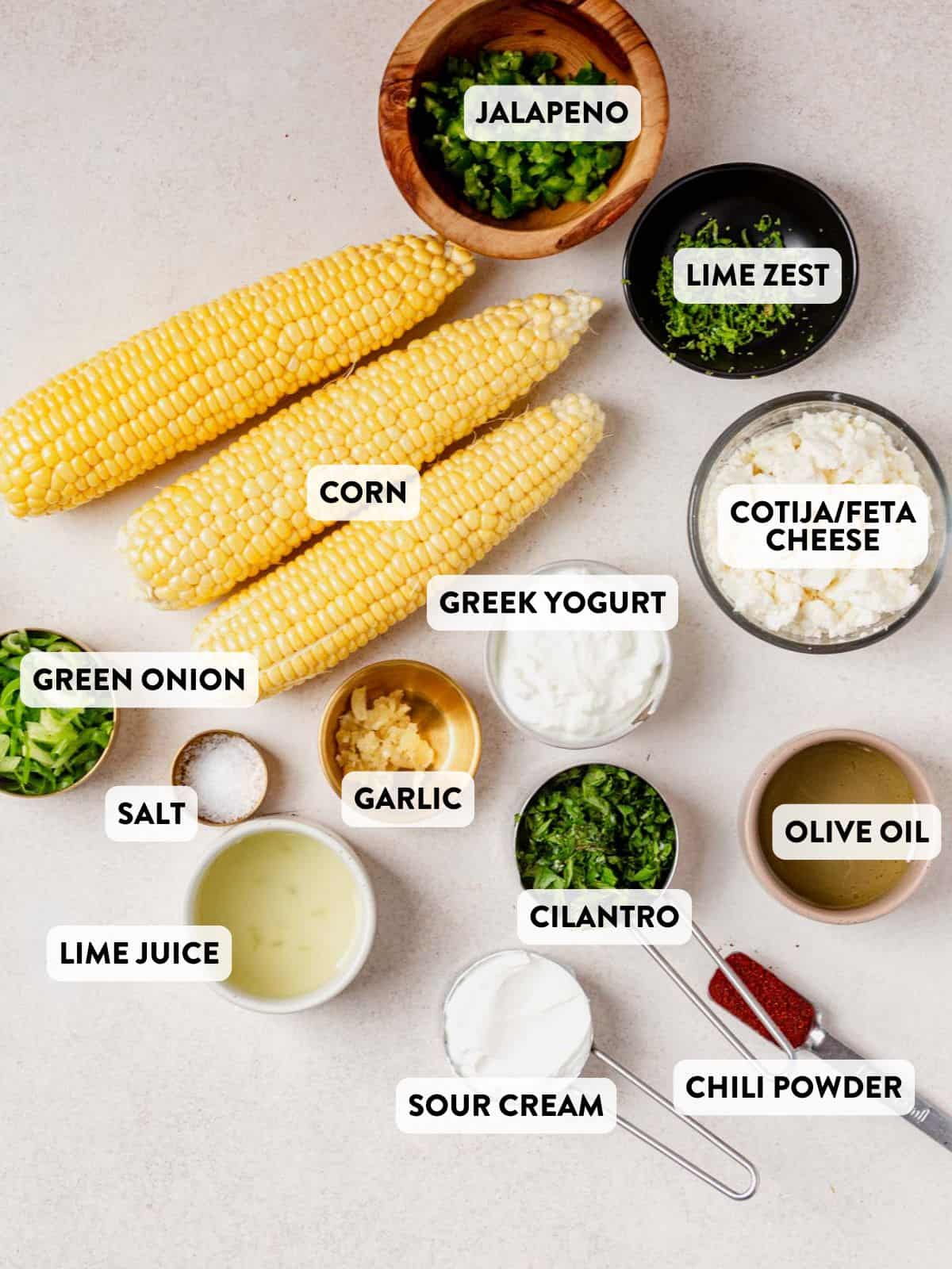 Overhead shot of labeled ingredients for Mexican street corn salad, including corn, lime juice, Greek yogurt, sour cream, jalapeño, cotija cheese, cilantro, and spices.