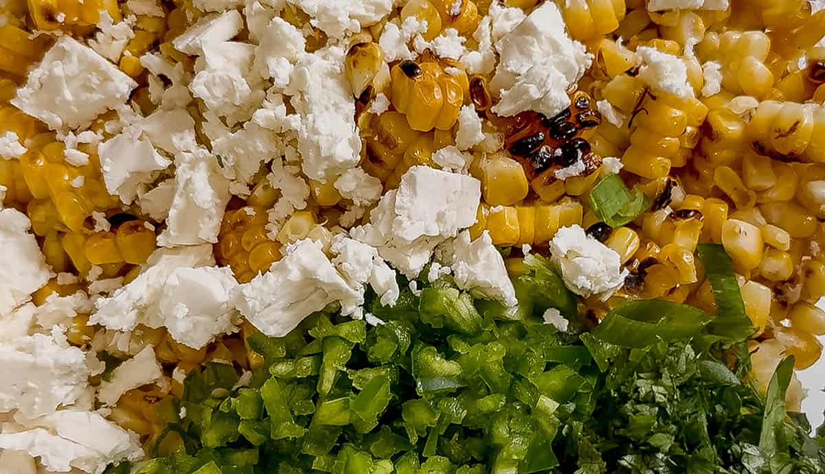 Close-up of grilled corn kernels topped with crumbled feta cheese, diced jalapeño, and chopped cilantro in a mixing bowl.