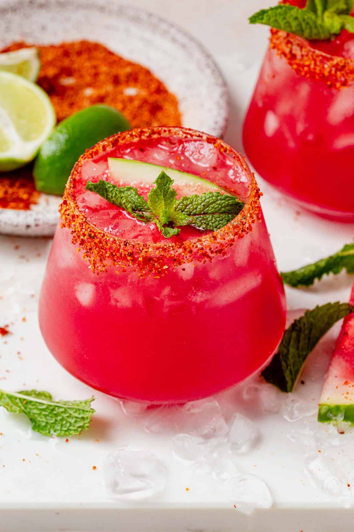 Bright pink watermelon vodka cocktail with ice, mint, and a watermelon slice, served in a Tajin-rimmed glass.