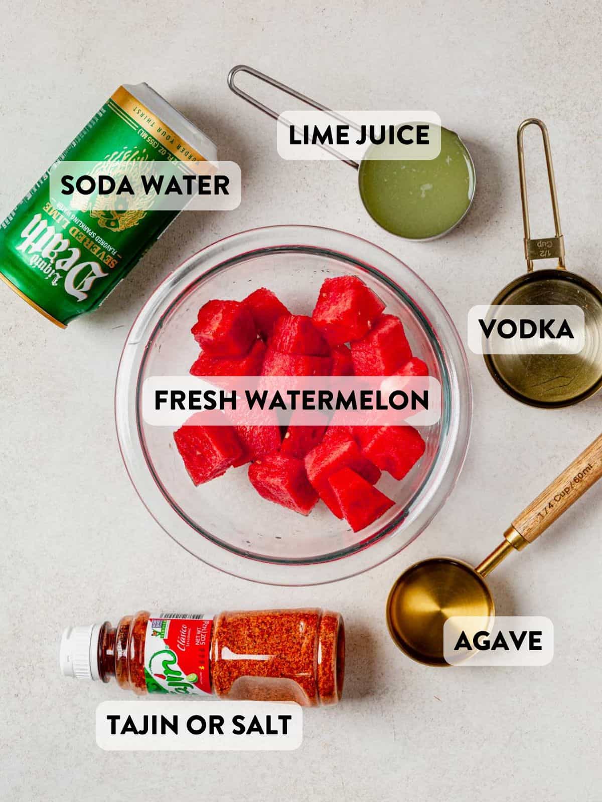 Overhead view of labeled ingredients for watermelon vodka cocktail including fresh watermelon, vodka, lime juice, agave, soda water, and Tajin.