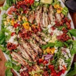 Overhead view of a colorful grilled chicken salad on a white platter with sliced grilled chicken breasts, charred corn, cherry tomatoes, avocado slices, red onion, crumbled feta, crispy bacon, and fresh greens.