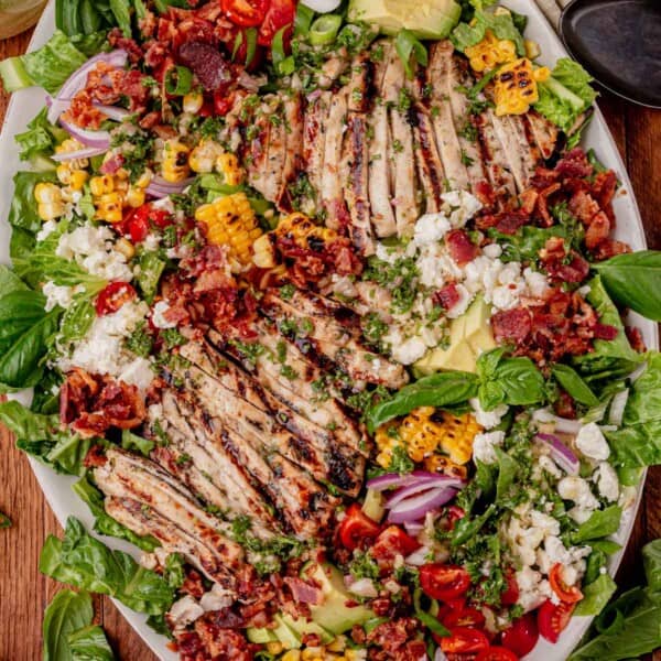 Overhead view of a colorful grilled chicken salad on a white platter with sliced grilled chicken breasts, charred corn, cherry tomatoes, avocado slices, red onion, crumbled feta, crispy bacon, and fresh greens.