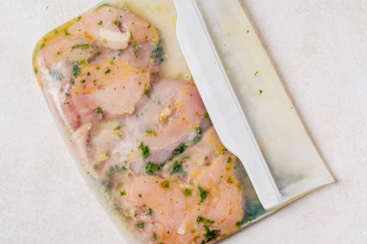 Raw chicken breasts marinating in a honey lemon herb mixture inside a reusable silicone bag on a light surface.
