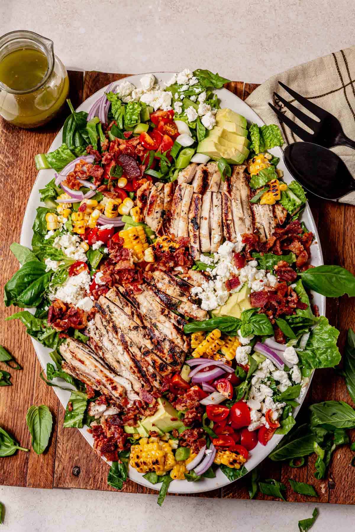 Large white platter filled with grilled chicken salad topped with charred corn, avocado slices, cherry tomatoes, red onion, crumbled feta, crispy bacon, and fresh greens, with a jar of dressing and serving utensils on the side.