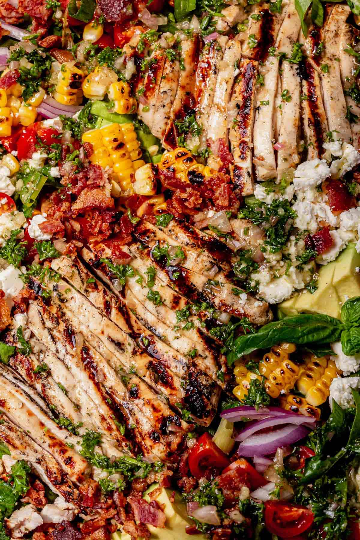 Close-up of sliced grilled chicken on a salad with charred corn, avocado, cherry tomatoes, red onion, crumbled feta, crispy bacon, and fresh herbs.