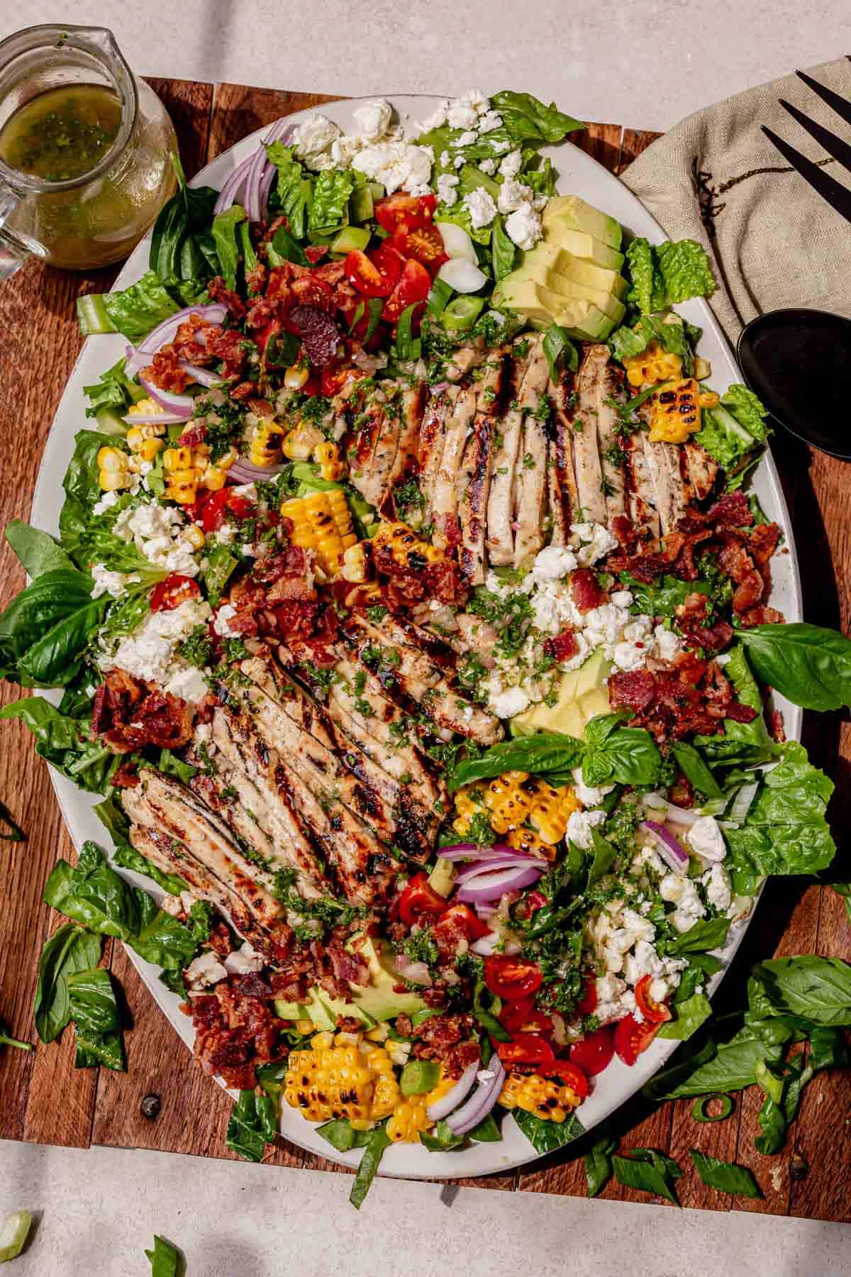 Platter of grilled chicken salad topped with charred corn, avocado, cherry tomatoes, red onion, crumbled feta, and crispy bacon, drizzled with fresh herb and honey lemon dressing.