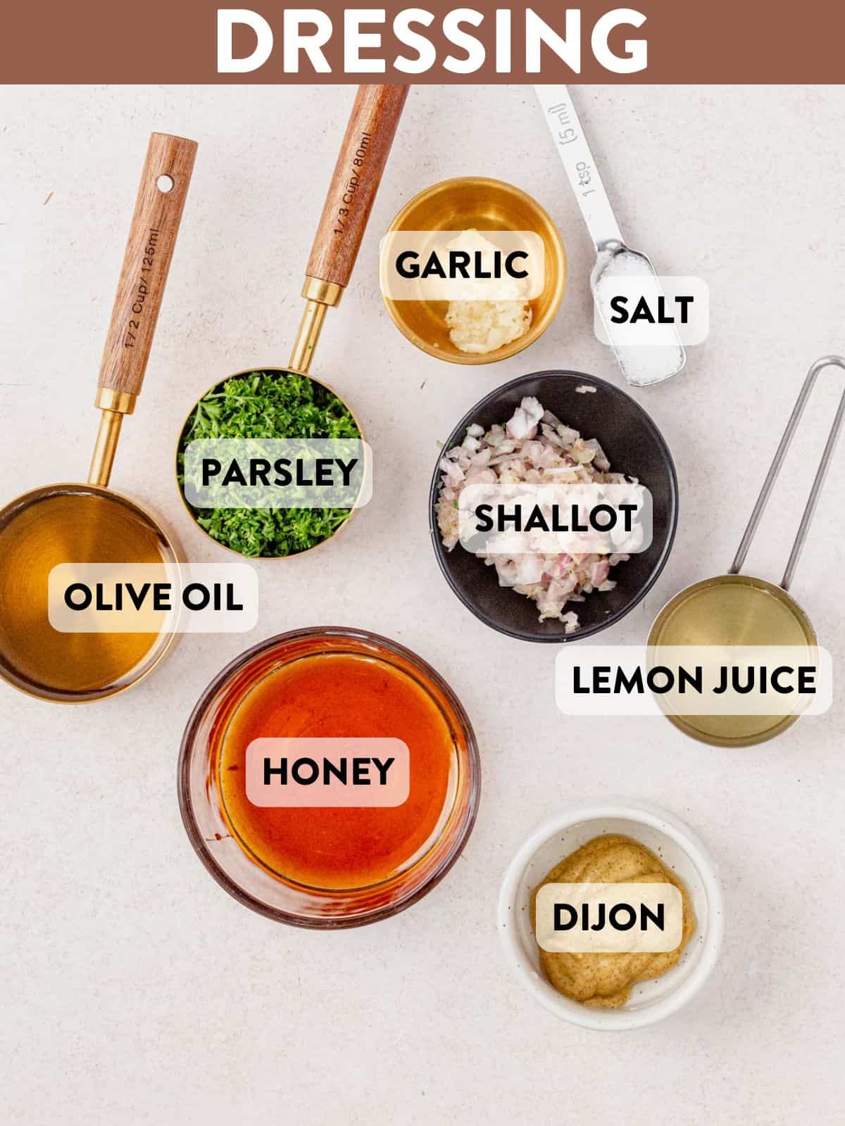 Overhead view of labeled ingredients for honey lemon dressing, including olive oil, parsley, garlic, salt, shallot, lemon juice, honey, and Dijon mustard, arranged on a light surface.