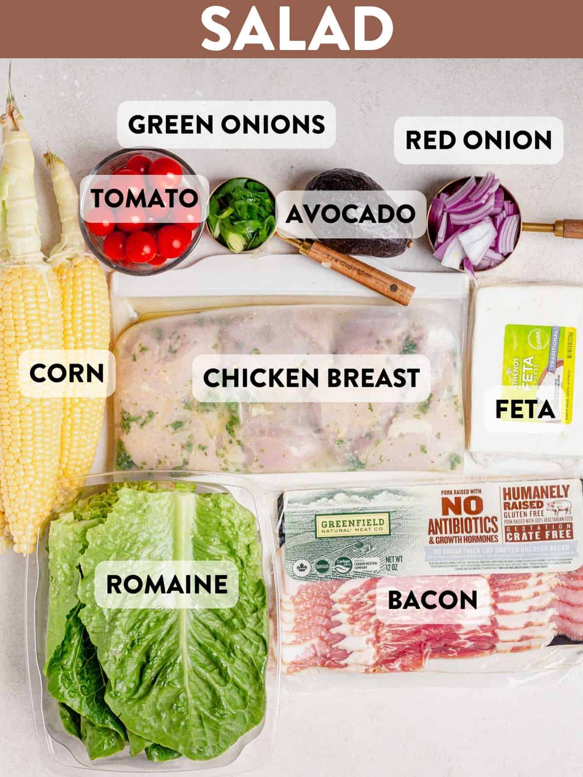 Overhead view of labeled ingredients for grilled chicken salad, including fresh corn, romaine lettuce, cherry tomatoes, green onions, avocado, red onion, feta cheese, bacon, and marinated chicken breast, arranged on a light surface.