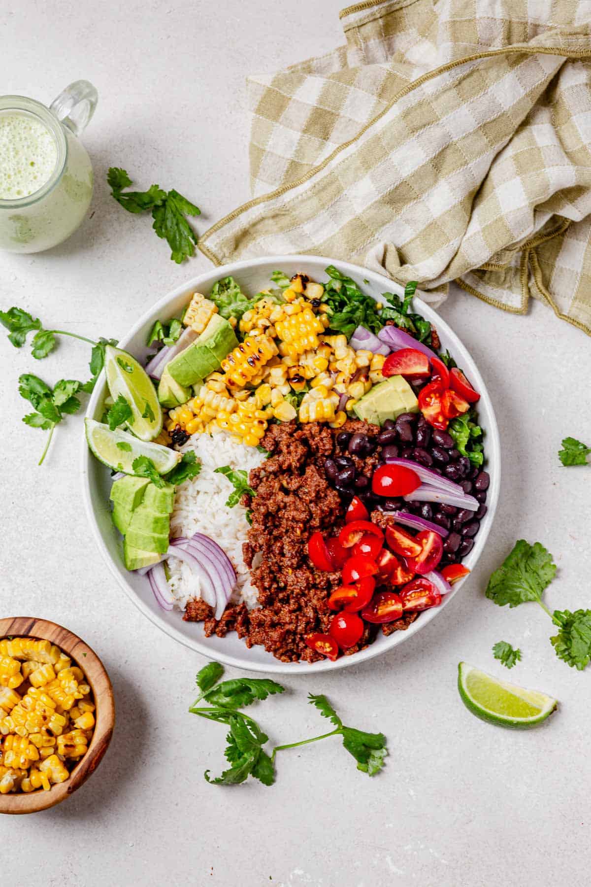 a taco bowl filled with white rice, ground beef, tomatoes, black beans, and surrounded by fresh cilantro and a side of creamy dressing.