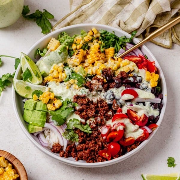 taco bowls in a white bowl with seasoned ground beef, rice, avocado, tomato, onion, corn, black beans, and dressing.