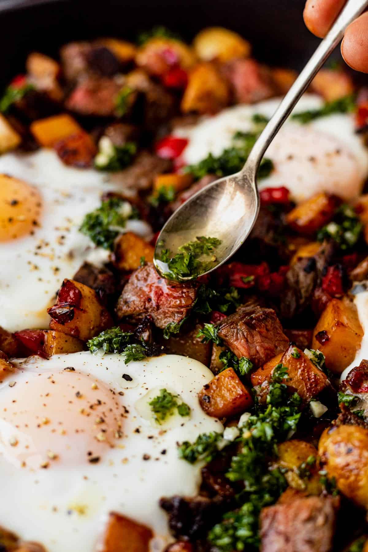 Spoon pouring chimichurri sauce over steak has with eggs and hashbrowns