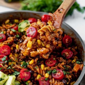 A wooden spoon scoops a hearty skillet meal reminiscent of taco bowls, featuring pasta, ground meat, corn, black beans, diced tomatoes, avocado, and fresh herbs. A blurred avocado and herbs are in the background.