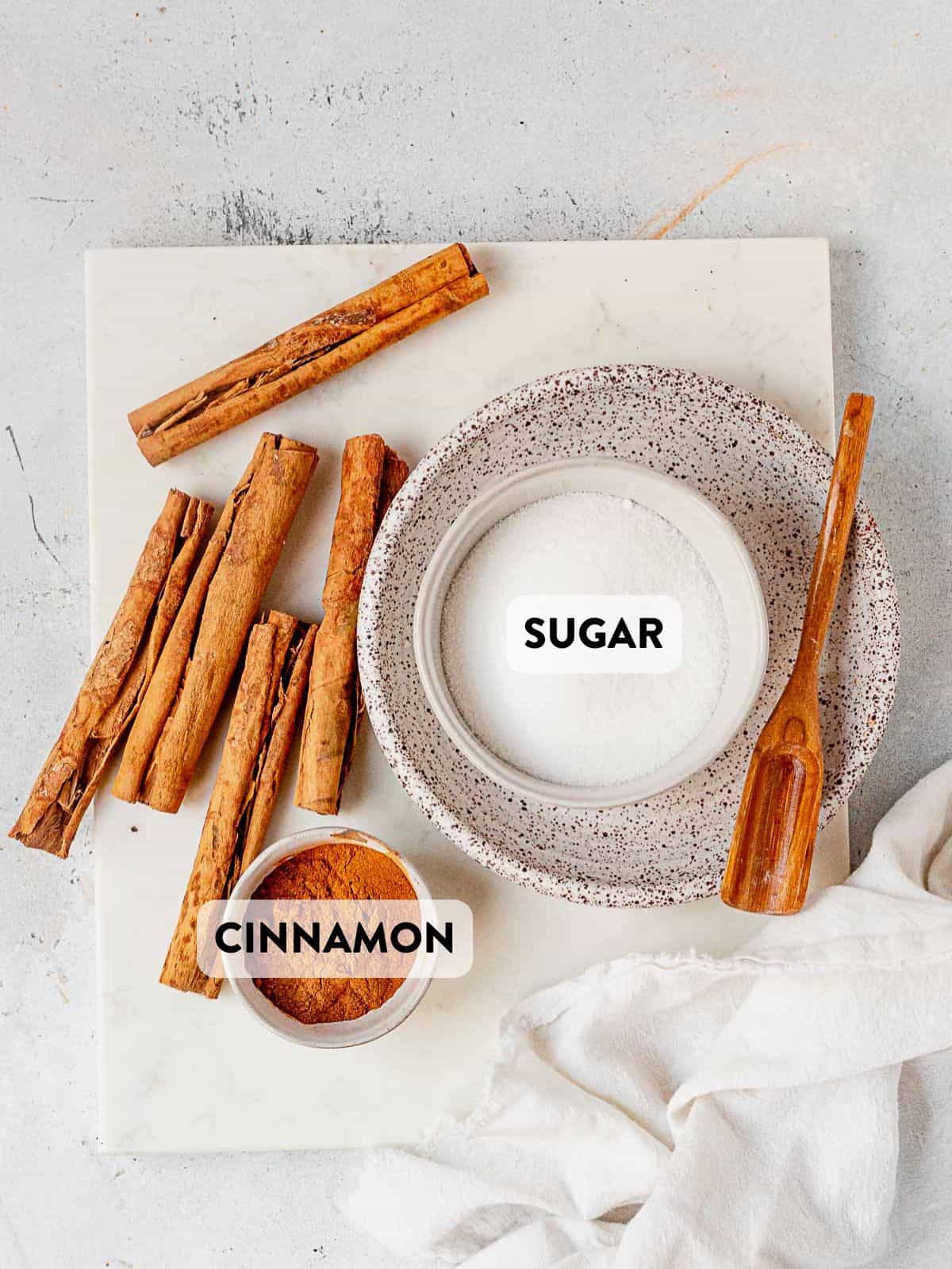 ingredients for cinnamon sugar mixture laid out on a counter in tiny bowls and cinnamon sticks next to them