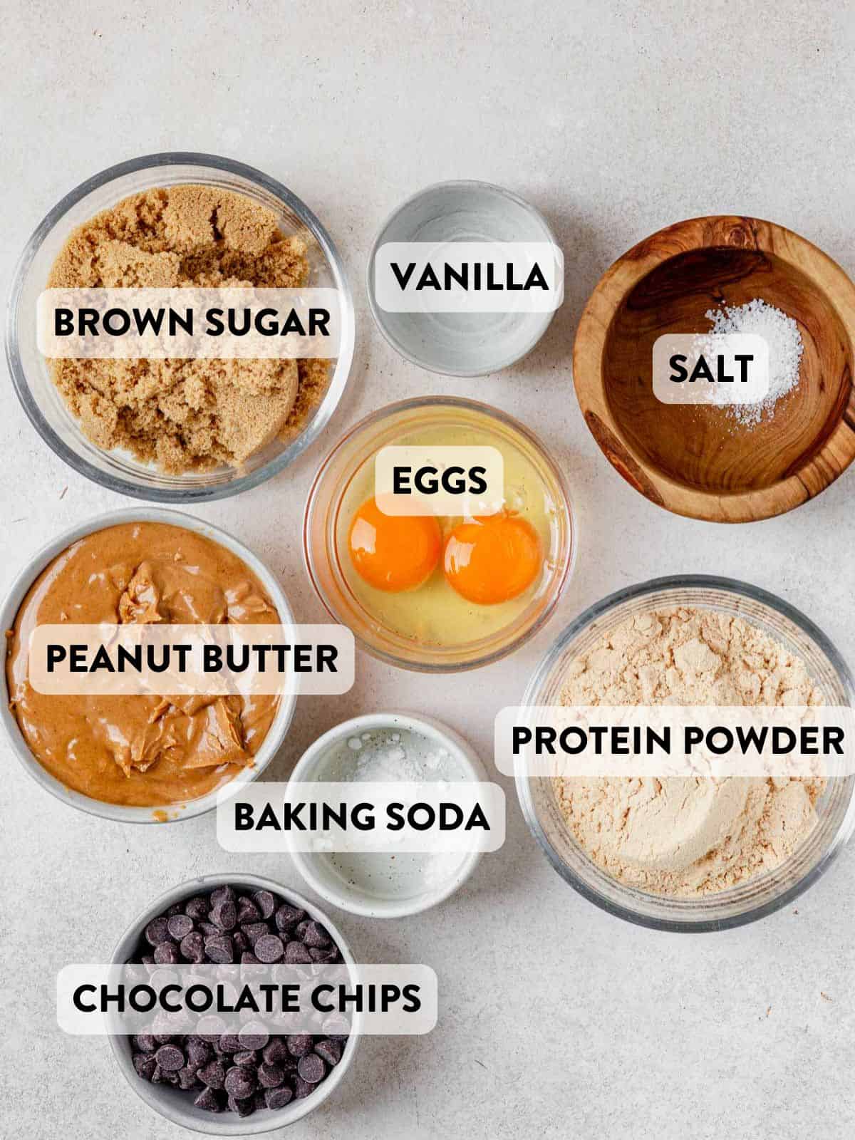 sugar, peanut butter, protein powder, vanilla, chocolate chips, and baking soda on a counter.