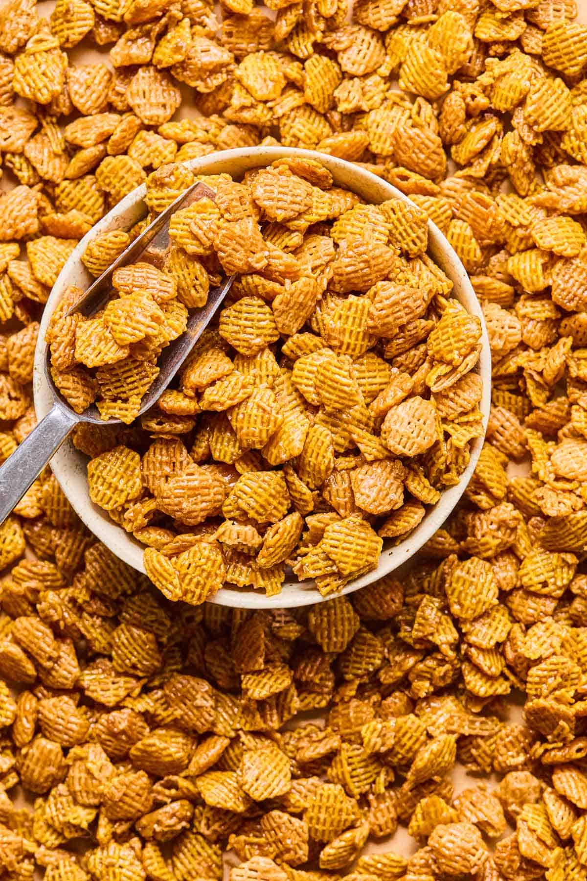 a metal scoop dipped into a bowl of crispix mix that spills out onto the counter around the bowl