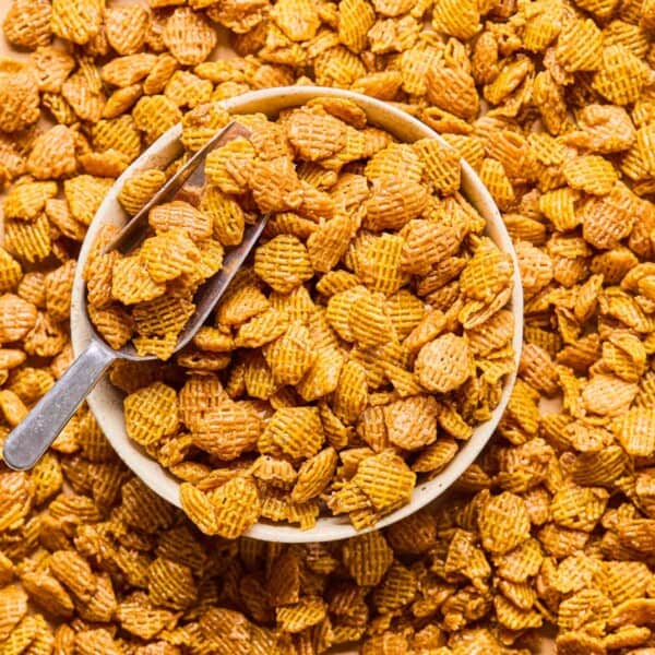 a metal scoop dipped into a bowl of crispix mix that spills out onto the counter around the bowl