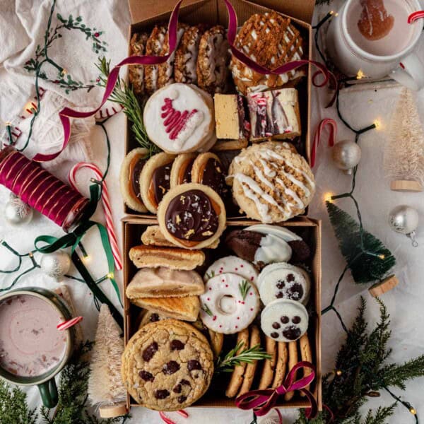 a christmas cookie box filled with holiday cookies and surrounded by lights and hot cocoa.